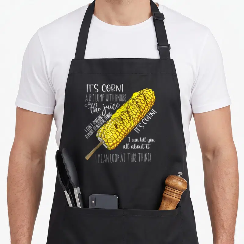 Lump corn apron - Cartoon yellow corn on a stick with white text reading It's Corn A Big Lump With Knobs — shown on a black cotton canvas apron against a white background