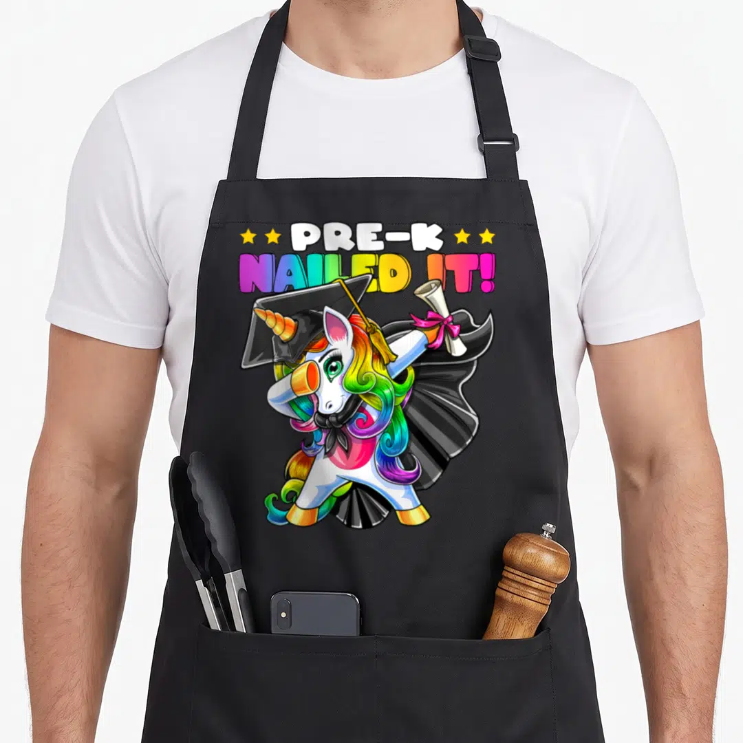 Cap unicorn apron - Dabbing unicorn in cap gown holding diploma with PRE-K NAILED IT! text and yellow stars — shown on a black cotton canvas apron against a white background