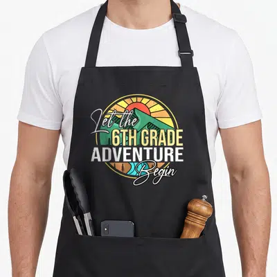 Begin school teacher apron - Retro circular mountain sunset with Let the 6th Grade Adventure Begin text in mixed fonts — shown on a black cotton canvas apron against a white background