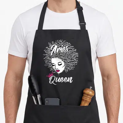 Zodiac afro birthday apron - Aries Queen text with woman's face and pink crown formed by white words — shown on a black cotton canvas apron against a white background