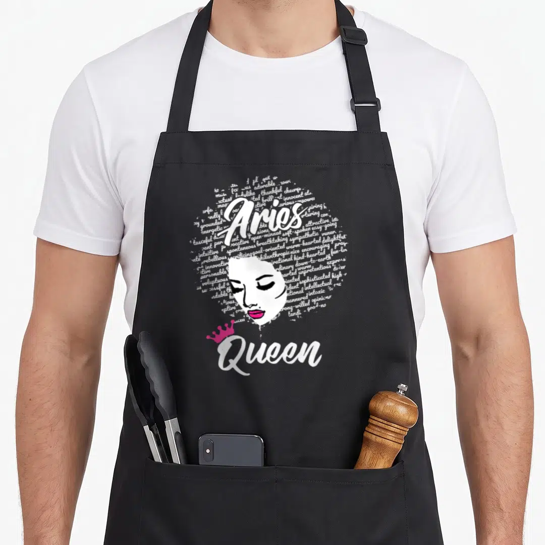 Zodiac afro birthday apron - Aries Queen text with woman's face and pink crown formed by white words — shown on a black cotton canvas apron against a white background