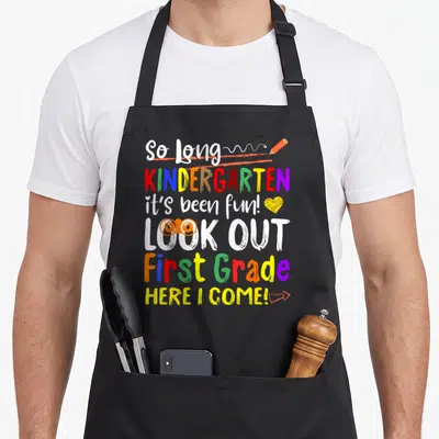 Kindergarten grade apron - Colorful stacked text reading So Long Kindergarten First Grade with pencil heart and glasses — shown on a black cotton canvas apron against a white background