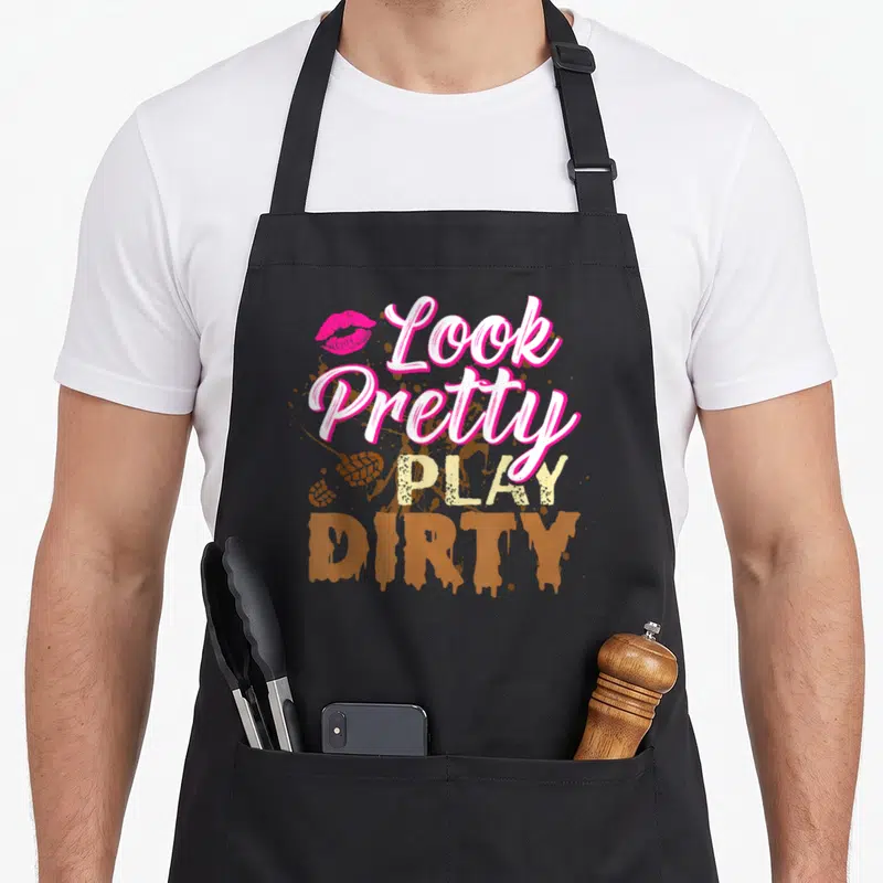 Pretty the dirty apron - Stacked Look Pretty Play Dirty text with pink lips and brown mud splatters — shown on a black cotton canvas apron against a white background