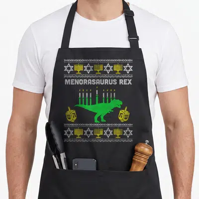 Menorasaurus jewish apron - Pixel art green T-Rex menorah with yellow dreidels and Stars of David border — shown on a black cotton canvas apron against a white background