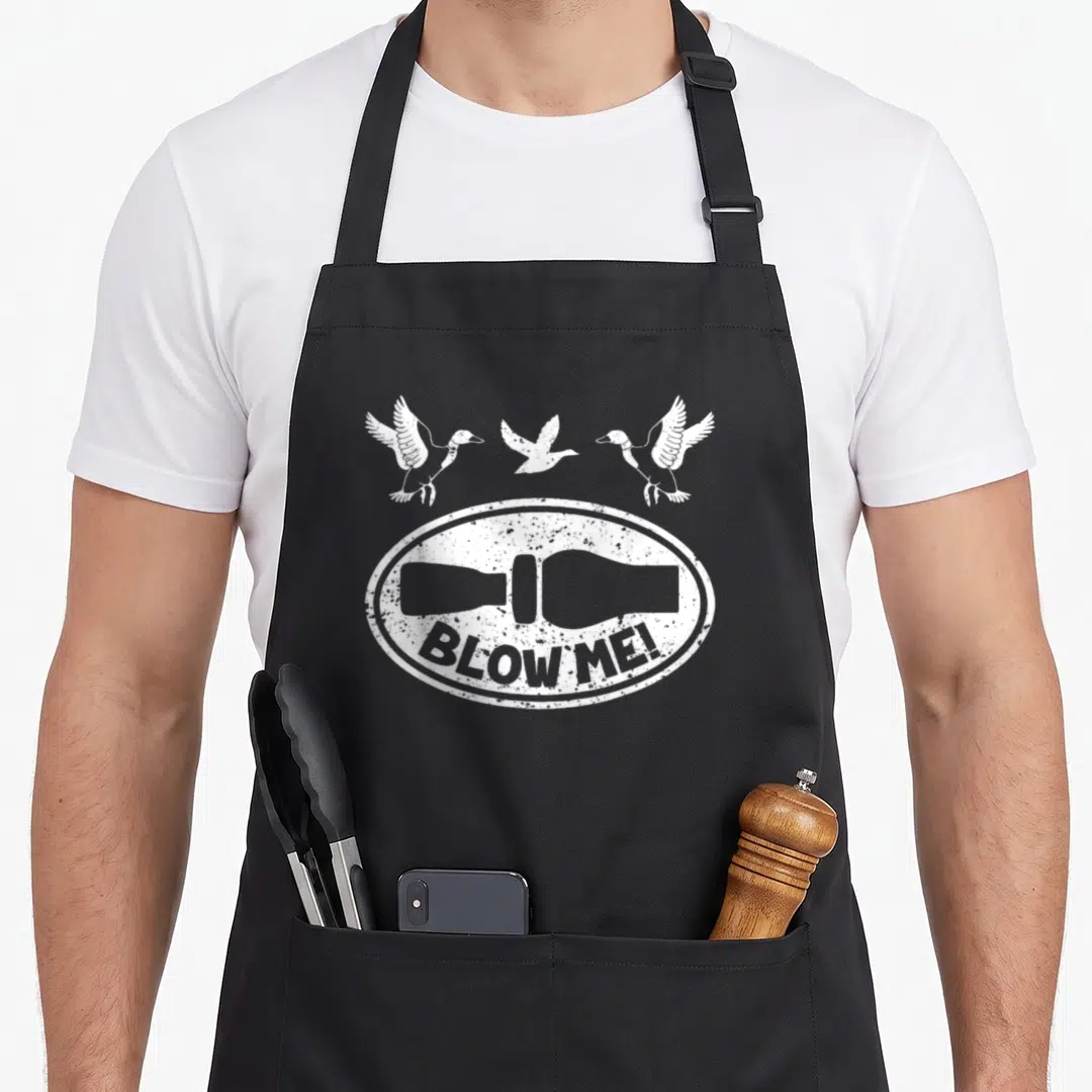 Duck hunter apron - White distressed duck call silhouette with BLOW ME! text and three flying ducks above an oval frame — shown on a black cotton canvas apron against a white background