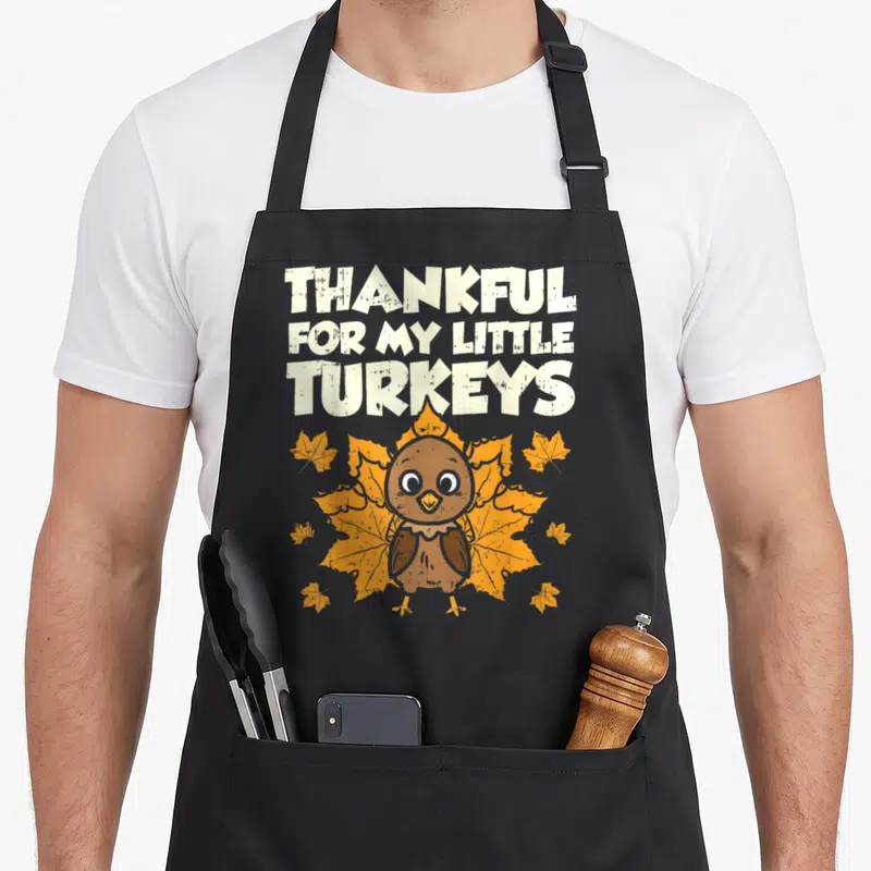 Thankful funny turkey apron - Cartoon turkey centered below stacked text over a large orange leaf with smaller — shown on a black cotton canvas apron against a white background