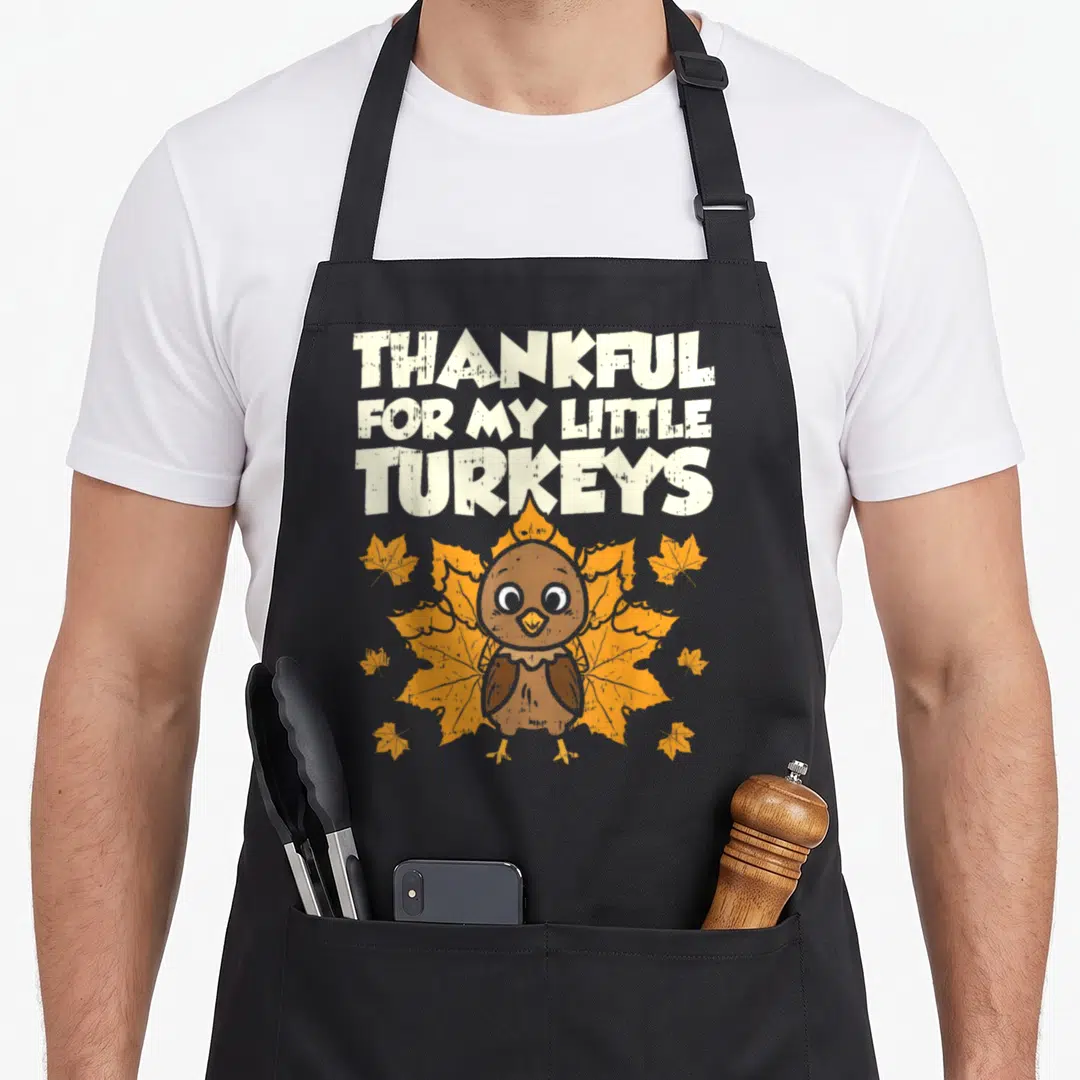 Thankful funny turkey apron - Cartoon turkey centered below stacked text over a large orange leaf with smaller — shown on a black cotton canvas apron against a white background