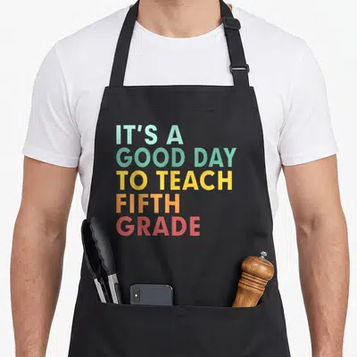 School teach teacher apron - Stacked text reading IT'S A GOOD DAY TO TEACH FIFTH GRADE in rainbow colors — shown on a black cotton canvas apron against a white background