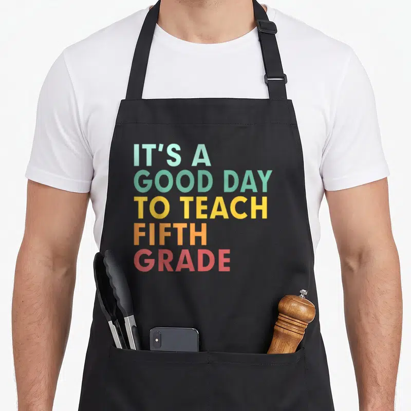 School teach teacher apron - Stacked text reading IT'S A GOOD DAY TO TEACH FIFTH GRADE in rainbow colors — shown on a black cotton canvas apron against a white background