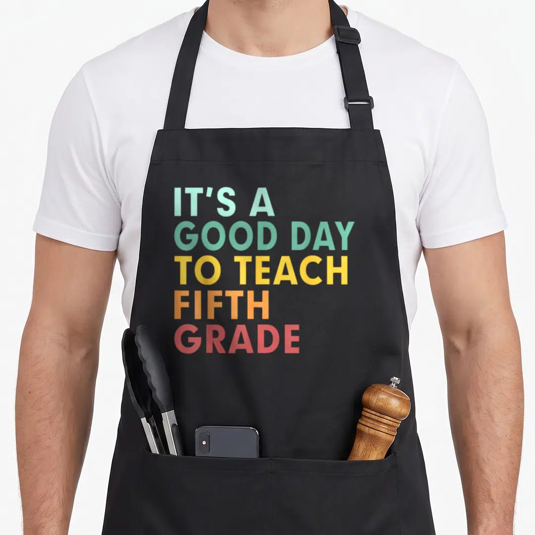 School teach teacher apron - Stacked text reading IT'S A GOOD DAY TO TEACH FIFTH GRADE in rainbow colors — shown on a black cotton canvas apron against a white background