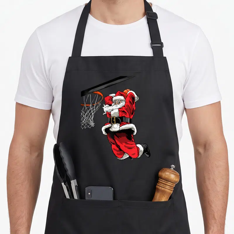 Basketball christmas santa apron - Cartoon Santa Claus jumping to dunk a basketball into a hoop in red and black — shown on a black cotton canvas apron against a white background