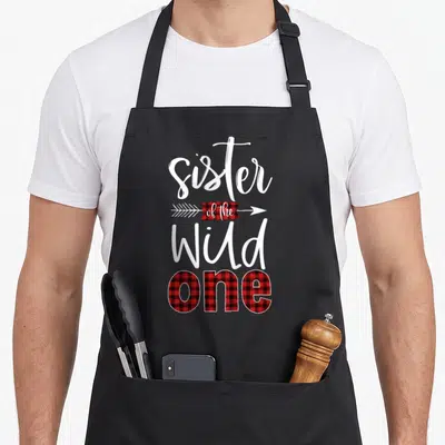 Sister the plaid apron - Vertical text sister of the Wild one with red black buffalo plaid arrow and filled letters — shown on a black cotton canvas apron against a white background