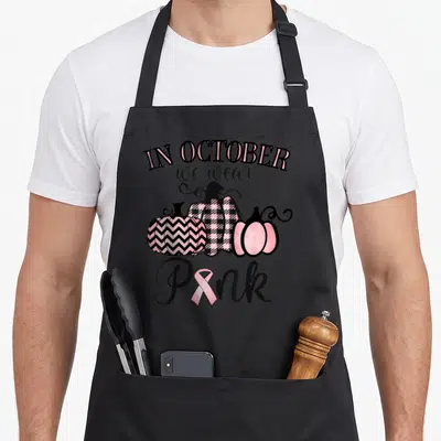 October breast cancer awareness aprons - Three patterned pumpkins below text reading IN OCTOBER we wear Pink — shown on a black cotton canvas apron against a white background