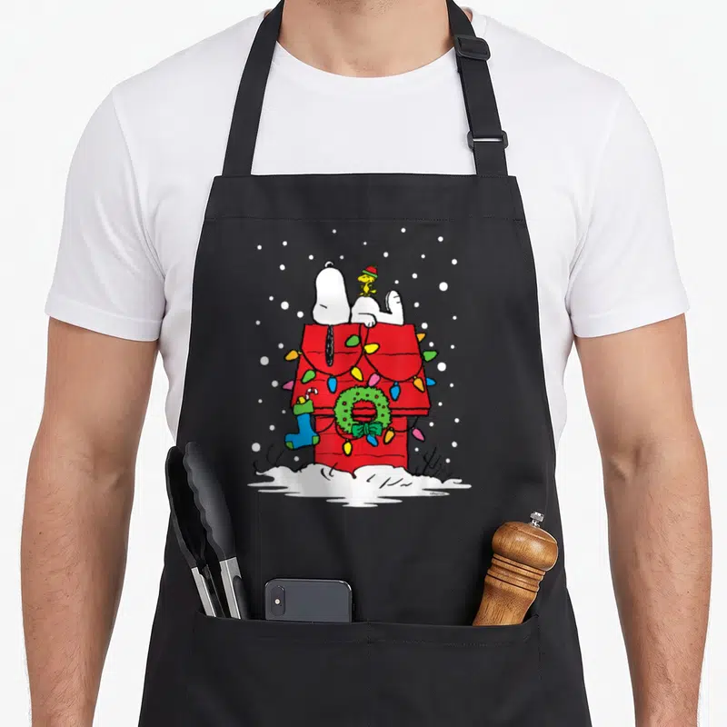 Woodstock holiday apron - Snoopy and Woodstock on a red doghouse with lights, wreath, stocking, and snow — shown on a black cotton canvas apron against a white background