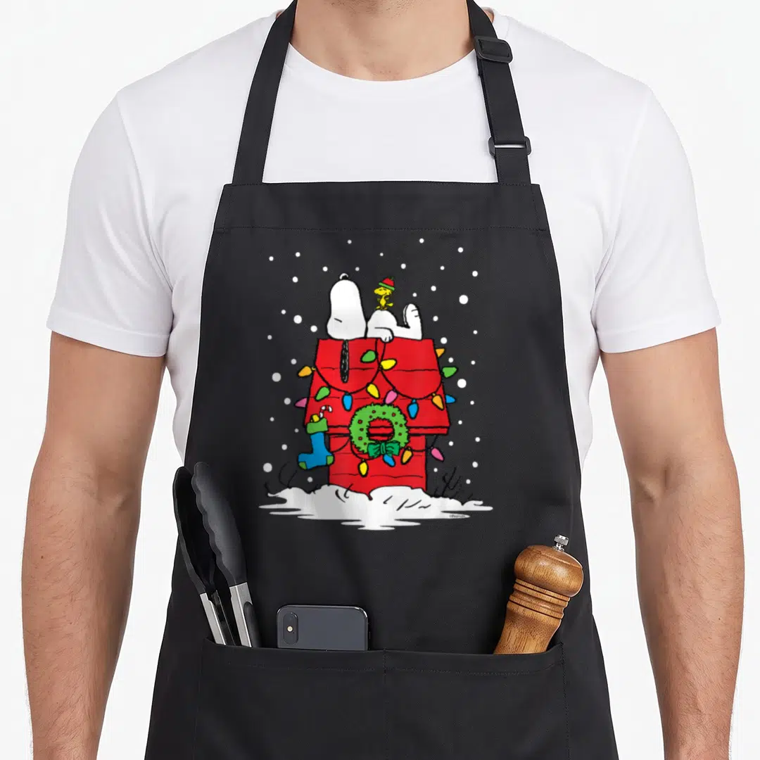 Woodstock holiday apron - Snoopy and Woodstock on a red doghouse with lights, wreath, stocking, and snow — shown on a black cotton canvas apron against a white background