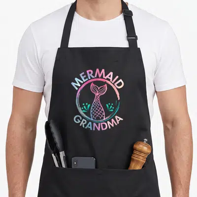 Mermaid birthday grandma in apron - Cartoon mermaid tail in pink blue purple with arched text MERMAID GRANDMA — shown on a black cotton canvas apron against a white background
