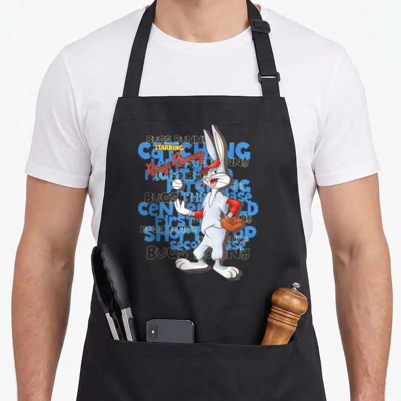 Baseball bunny apron - Bugs Bunny standing in front of repeated BUGS BUNNY text in grey, white, blue, red, yellow — shown on a black cotton canvas apron against a white background