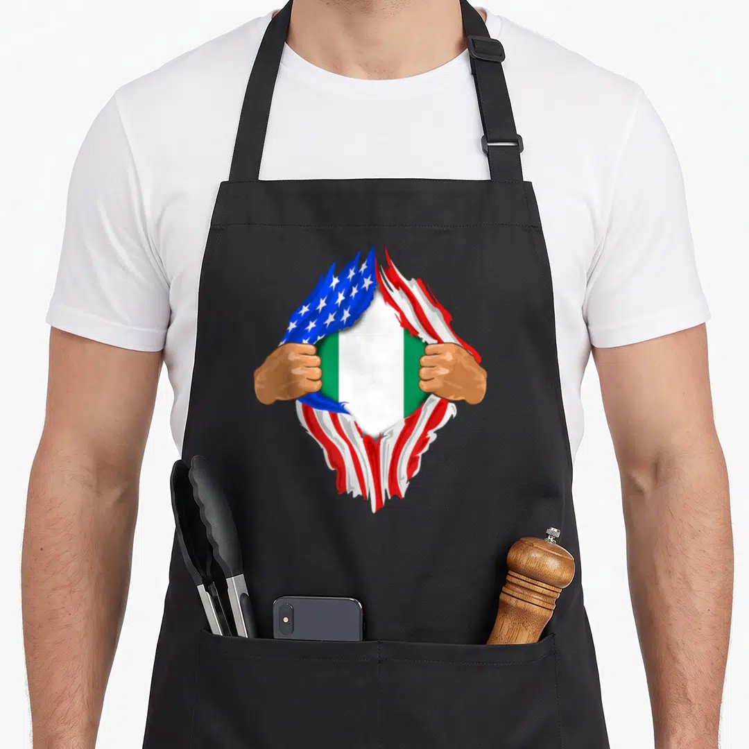 Nigerian blood apron - Two brown hands tearing a US flag to reveal the Nigerian green white and red flag — shown on a black cotton canvas apron against a white background