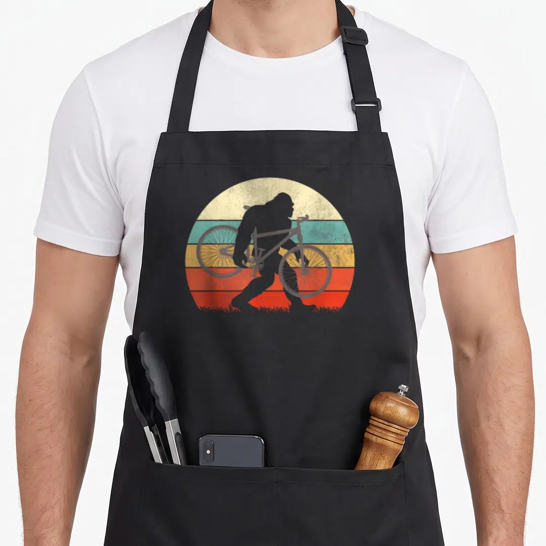 Bike bigfoot apron - Retro gorilla silhouette carrying a bike against cream teal yellow orange and red striped sun — shown on a black cotton canvas apron against a white background