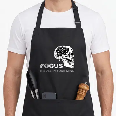 Focus darts addict apron - Distressed skull with dartboard inside cranium and stacked text FOCUS IT'S ALL IN YOUR MIND — shown on a black cotton canvas apron against a white background