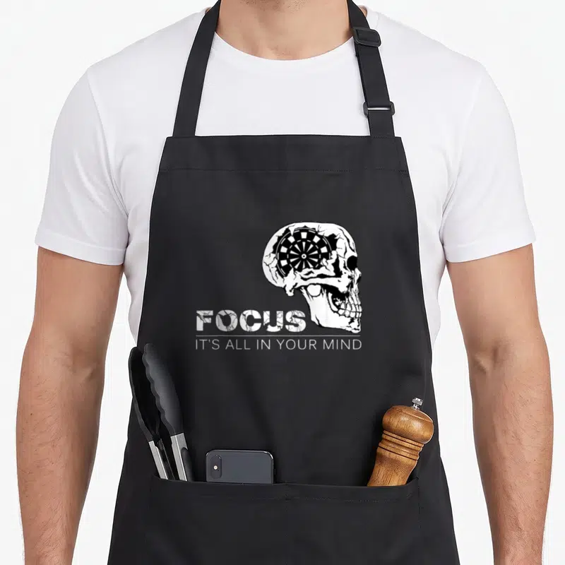 Focus darts addict apron - Distressed skull with dartboard inside cranium and stacked text FOCUS IT'S ALL IN YOUR MIND — shown on a black cotton canvas apron against a white background