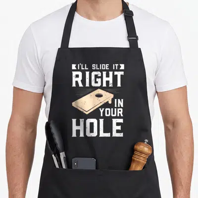 Ill game aprons - Beige cornhole board graphic with white distressed text reading I'll Slide It Right In Your Hole — shown on a black cotton canvas apron against a white background