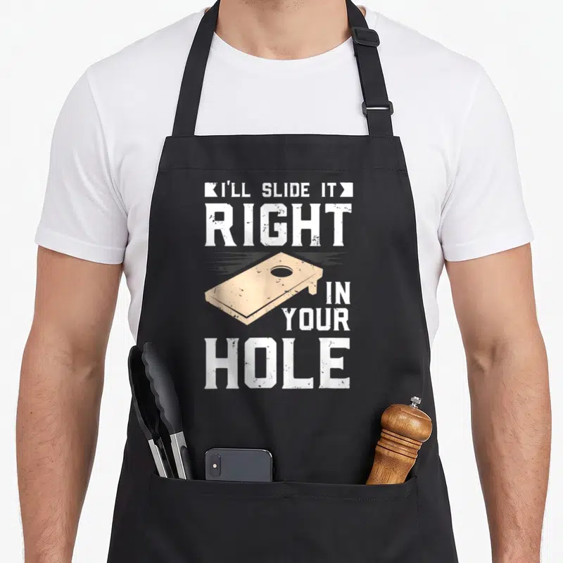 Ill game aprons - Beige cornhole board graphic with white distressed text reading I'll Slide It Right In Your Hole — shown on a black cotton canvas apron against a white background