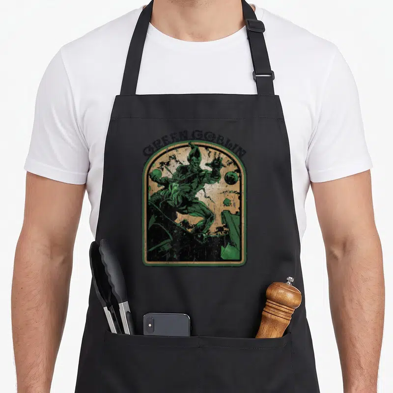 Comic goblin valley apron - Distressed Green Goblin on glider inside arched frame with arched text — shown on a black cotton canvas apron against a white background