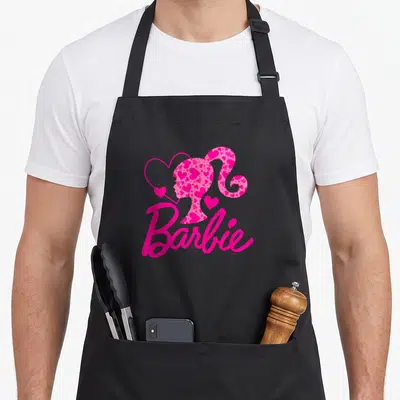 Funny crew apron - Pink Barbie silhouette filled with hearts and script logo — shown on a black cotton canvas apron against a white background