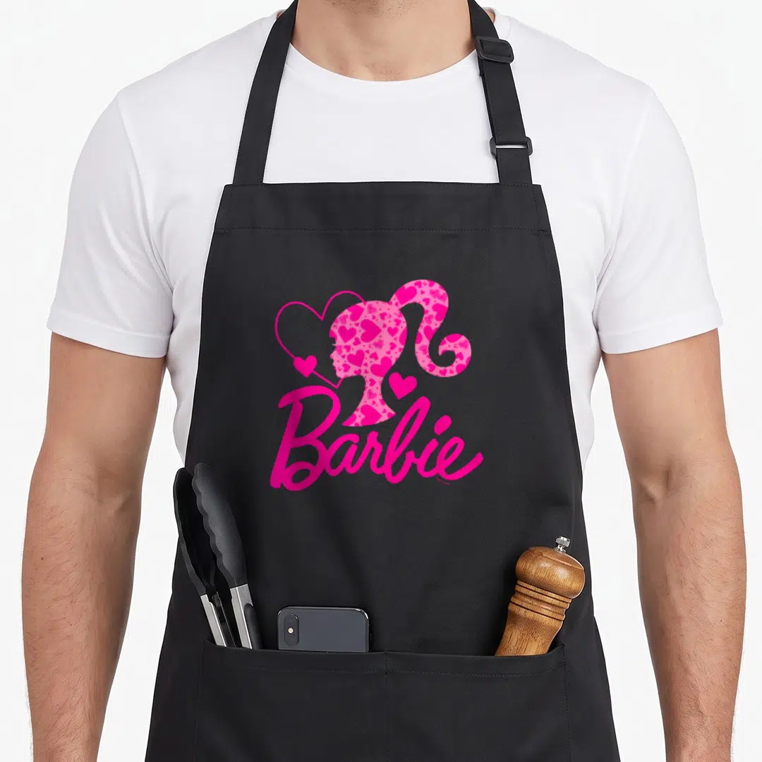Funny crew apron - Pink Barbie silhouette filled with hearts and script logo — shown on a black cotton canvas apron against a white background