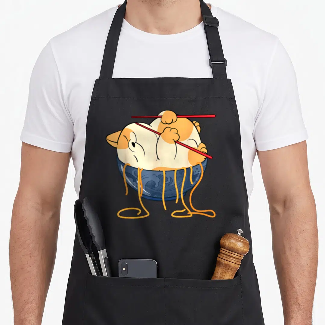 Clothe cat apron - Cartoon orange white cat curled in blue bowl holding red chopsticks with yellow noodles hanging down — shown on a black cotton canvas apron against a white background