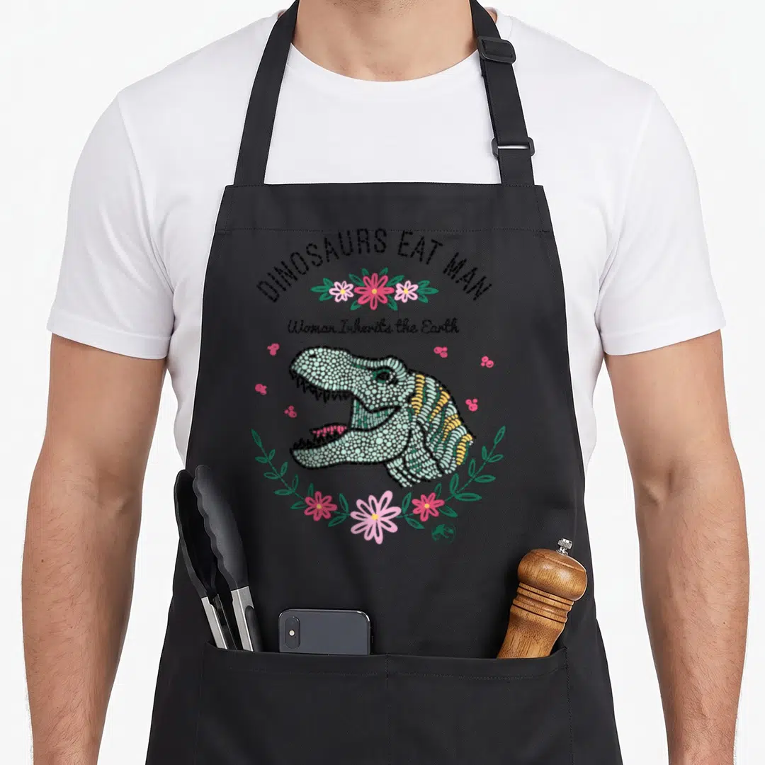 Dino earth apron - Green dinosaur head surrounded by pink flowers and black text reading DINOSAURS EAT MAN — shown on a black cotton canvas apron against a white background