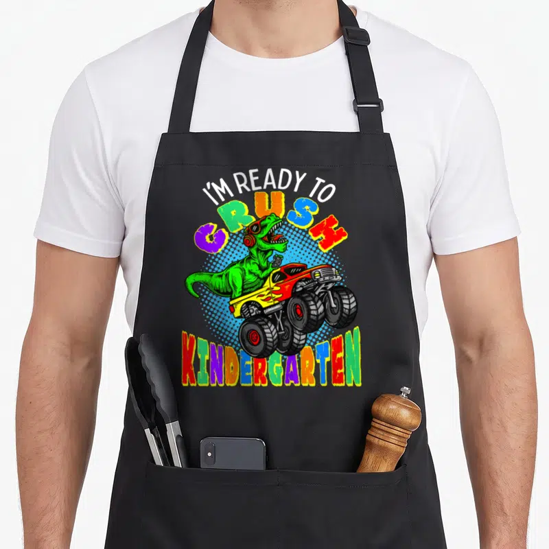 Kindergarten dinosaur truck apron - Green t-rex in red monster truck with colorful I'm Ready to Crush Kindergarten text — shown on a black cotton canvas apron against a white background
