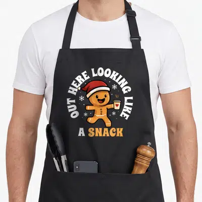 Snack christmas gingerbread apron - Cartoon gingerbread man in red hat holding coffee cup with OUT HERE LOOKING LIKE — shown on a black cotton canvas apron against a white background