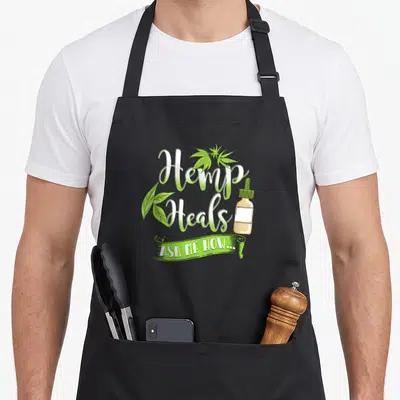 Heal hemp apron - Hemp Heals text, green leaves, tan dropper bottle, and ASK ME HOW banner — shown on a black cotton canvas apron against a white background