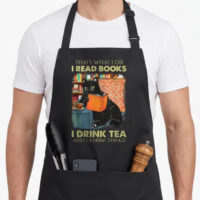 Read tea apron - Black cat reading orange book beside tea cup with humorous quote about knowing things — shown on a black cotton canvas apron against a white background