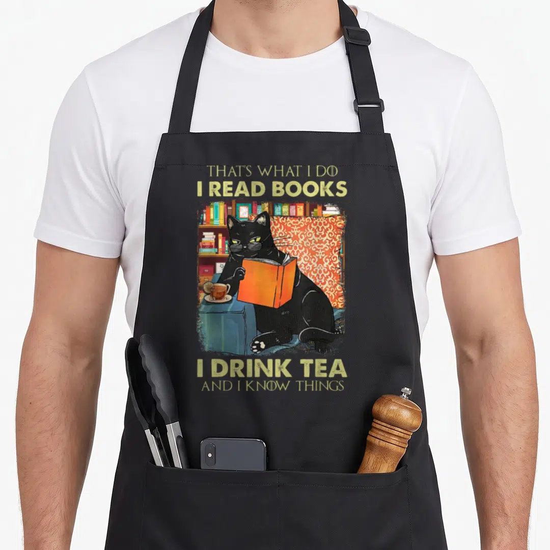 Read tea apron - Black cat reading orange book beside tea cup with humorous quote about knowing things — shown on a black cotton canvas apron against a white background