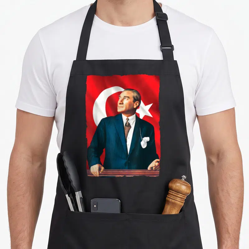 Mustafa founder apron - Man in blue suit at podium before red flag with white crescent and star — shown on a black cotton canvas apron against a white background