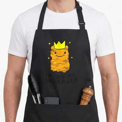 Funny beautiful chicken print apron - Smiling orange nugget wearing a yellow crown with four stars and curved text — shown on a black cotton canvas apron against a white background