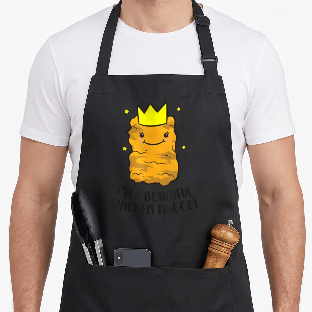 Funny beautiful chicken print apron - Smiling orange nugget wearing a yellow crown with four stars and curved text — shown on a black cotton canvas apron against a white background