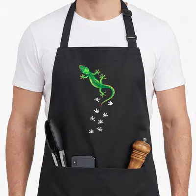 Lizard whisperer funny apron - Green gecko illustration with a trail of white footprint silhouettes descending — shown on a black cotton canvas apron against a white background