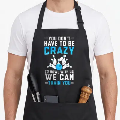 Funny bowler bowling player apron - Blue bowling ball knocking over six white pins with You Don't Have To Be Crazy text — shown on a black cotton canvas apron against a white background