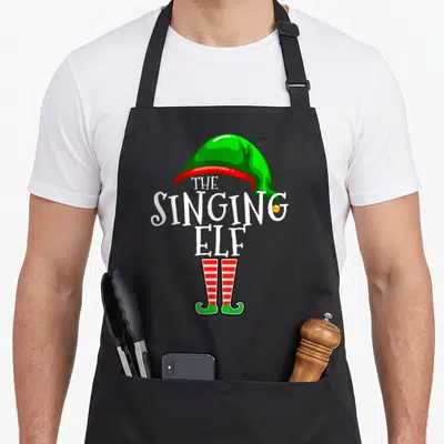 Singer christmas elf apron - White text forming elf body below green hat with red trim and striped legs — shown on a black cotton canvas apron against a white background