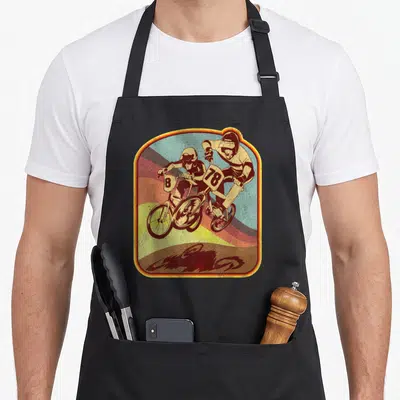Bmx 80s apron - Two motocross riders with numbers 8 and 78 on bikes against curved rainbow stripes in a retro frame — shown on a black cotton canvas apron against a white background