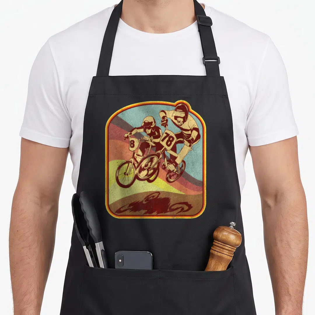Bmx 80s apron - Two motocross riders with numbers 8 and 78 on bikes against curved rainbow stripes in a retro frame — shown on a black cotton canvas apron against a white background