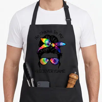Narcotic sobriety apron - Woman silhouette with rainbow tie-dye headband and sunglasses under No Shame In My text — shown on a black cotton canvas apron against a white background
