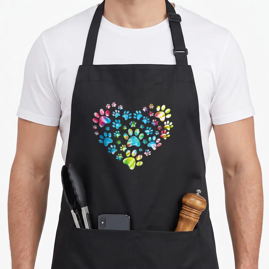 Tie paw print apron - Heart shape made of multicolored blue pink yellow green paw prints in watercolor style — shown on a black cotton canvas apron against a white background