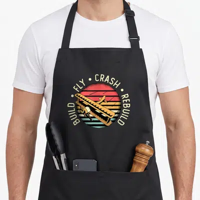 Build airplane apron - Retro sun with brown plane and Build Fly Crash Rebuild text — shown on a black cotton canvas apron against a white background