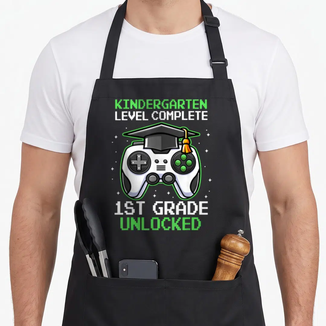 Kindergarten grade graduation apron - Game controller wearing a graduation cap with pixelated text reading level — shown on a black cotton canvas apron against a white background