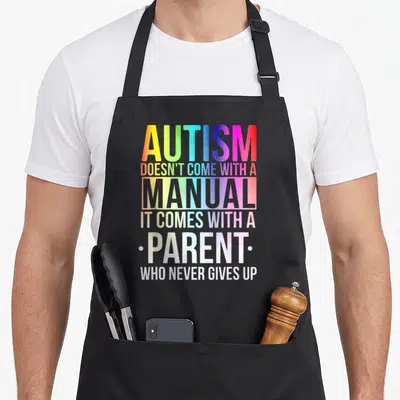 Come autism apron - Rainbow gradient text reading AUTISM DOESN'T COME WITH A MANUAL IT COMES WITH A PARENT WHO NEVER — shown on a black cotton canvas apron against a white background